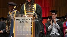 Central graduate and Honorary Fellowship Recipient Nia DaCosta stands at a podium on stage, wearing a cap and gown, addressing graduates