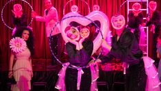 On a stage washed with pink lighting, an ensemble of actors in sequined outfits or ballgowns perform with hula-hoops and fluffy heart-shapes, pulling poses and looking through the holes in the props.