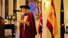 Nuria Espert wearing her Honorary Doctorate gown and hat stands in front of the Spanish Flag at the Residence of the Ambassador of Spain