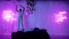 A performer dressed in white, stands on a bed, with their hands raised over their head. Behind them is a white curtain with pink design projection