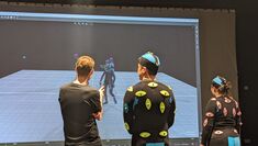 Two performers in MOCAP suits and the tutor stand facing large screen