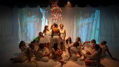 Performers dressed in white gather in a white bedroom set, with images projected on material behind them