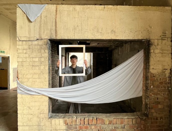 An actor appears within glassless brick window, half covered by draped material, holding a wooden frame to frame their head and shoulders