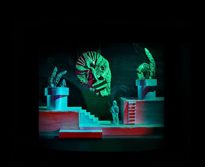 A red and green stage model of a character looking up at a mask and hands coming up out of the stage