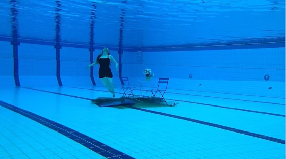A table and chairs under water in a blue swimming pool with one figure in bathing suit floating vertically