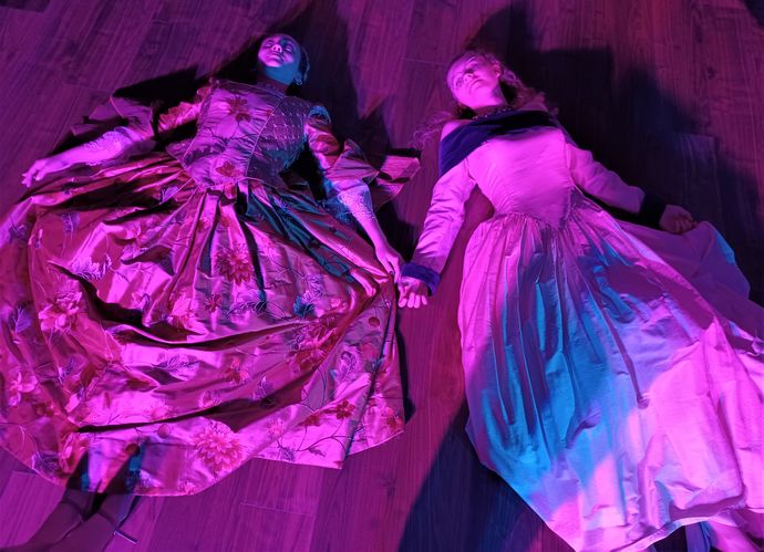 Two actors wearing dresses from Jacobean era, lie on a wooden floor, bathed in pink and blue light
