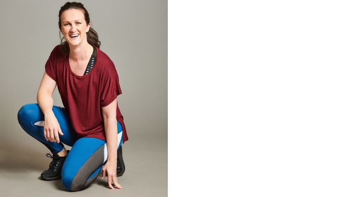 Portrait of Kim Wright wearing a red top and blue and black trousers. She is kneeling on one knee and smiling