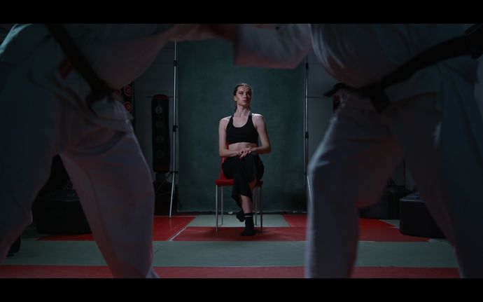 Dancer Bryony Bevan sits in focus in the background on a red chair. She looks to the corner, her legs are wrapped tightly crossed, and her chest and shoulders are open to the camera. Martial Artist Mick Garrod and Sensei Tony Newman are out of focus in the foreground, only their lower half is shown, their arms wrap tightly around each other’s shoulders.