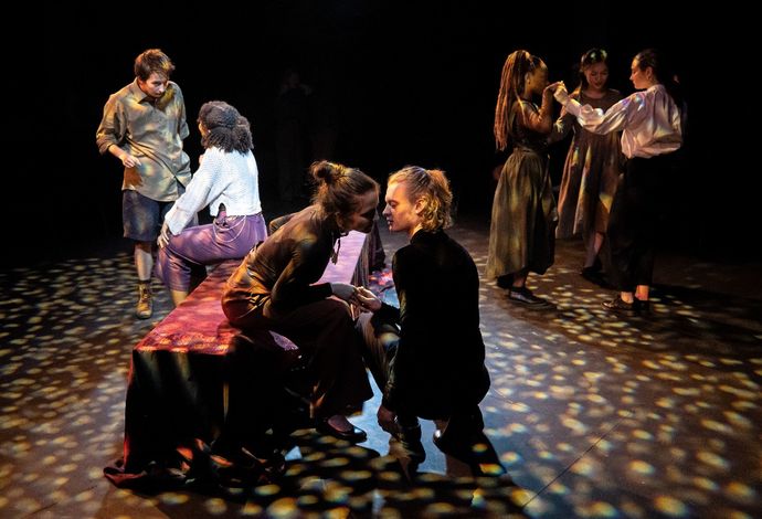 Students perform in a black box theatre space, there is a bench in the centre of the space, and the students are in groups of two and three under dappled light