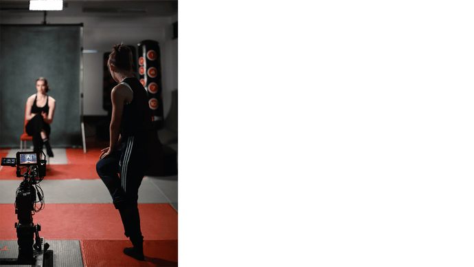 Bryony Bevan sits out of focus in the background on a red chair, inside a fight Dojo. The floor is red. Phoebe Stapleton stands in the foreground next to a film camera, she is movement directing Bryony. Both their legs are crossed and making a narrow position with their bodies.