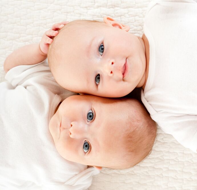 Two babies lie from opposite directions with their heads next to each other. They are wearing white tops against a white sheet